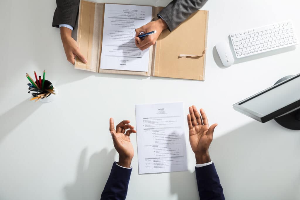 Elevated View Of Businessperson And Candidates Hand With Resume On White Desk